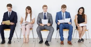 Professionally dressed people sit in a line looking at their resumes, in the hopes of landing a new job